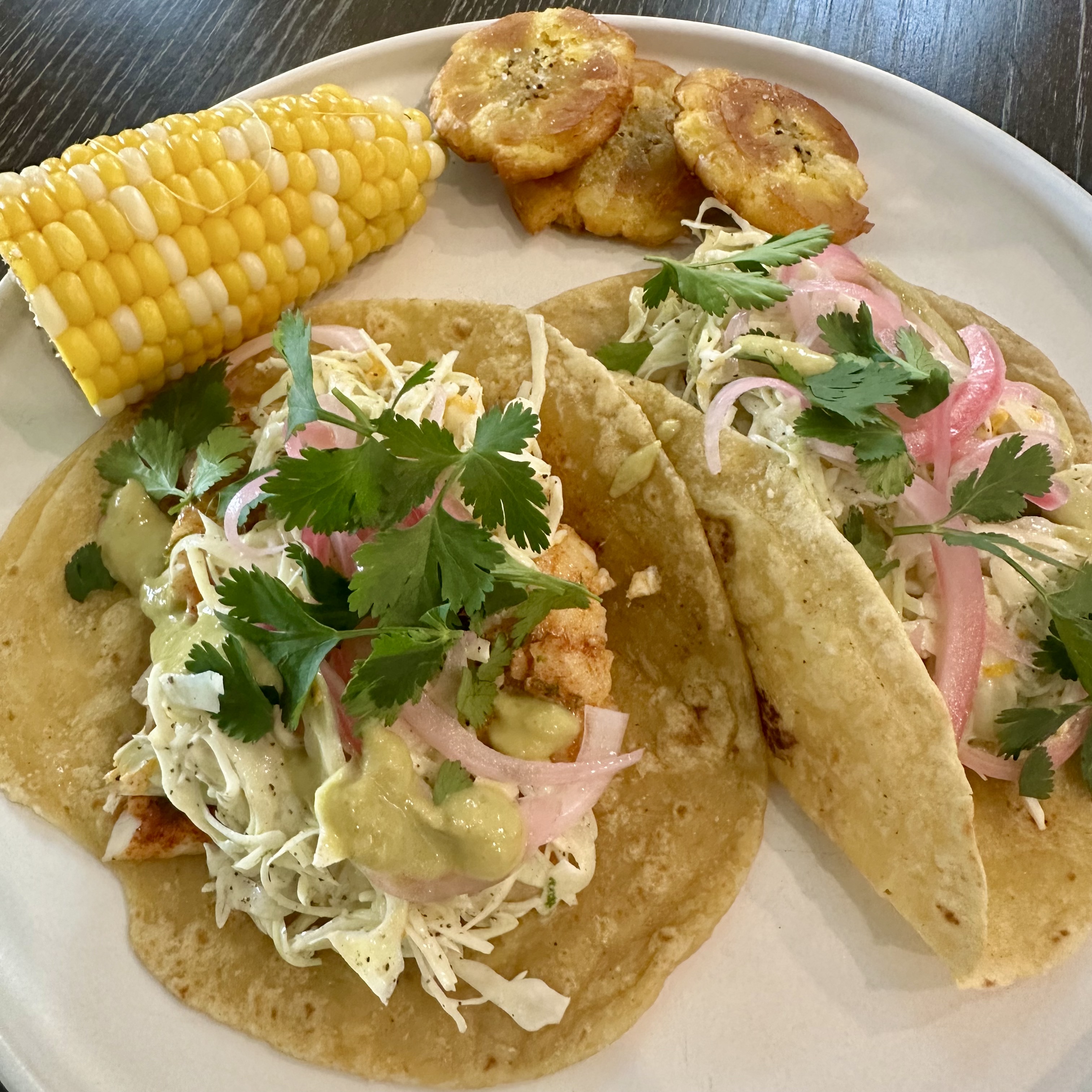 Rainbow trout fish tacos with a citrus slaw, pickled onions, jalapeno crema on mitad y mitad tortillas, served with tostones and roasted corn
